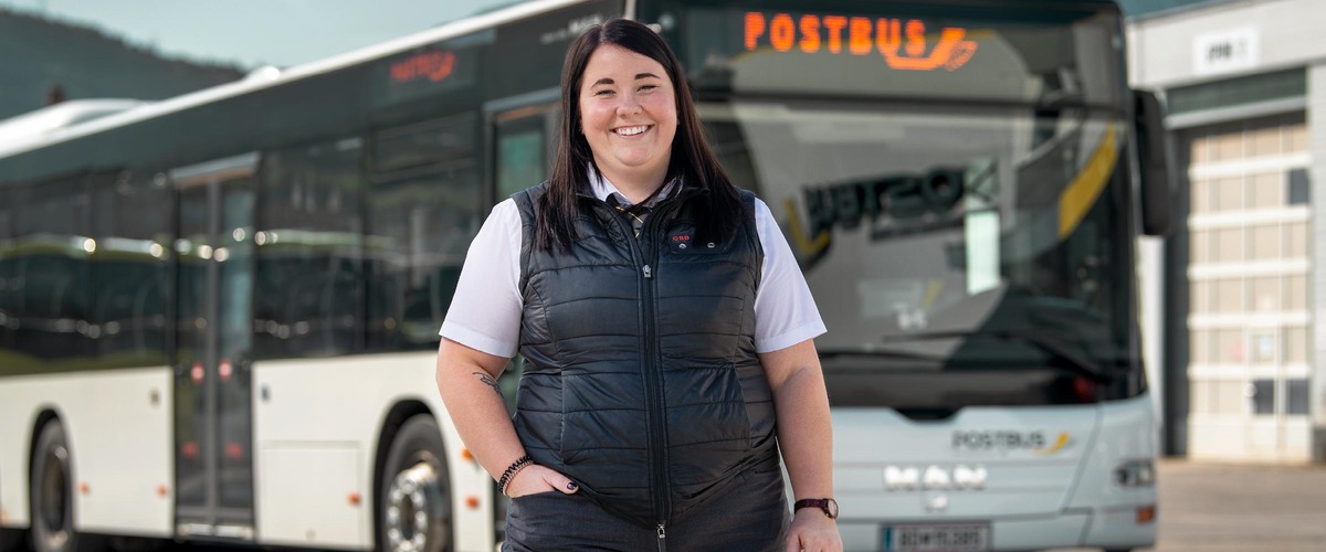 Frau in weißen Hand und schwarzen Gilet. Steht vor einem Postbus