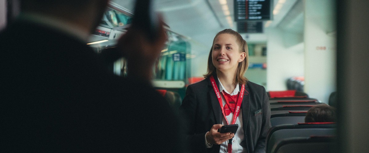 Eine weibliche Zugbegleiterin in einen Zug stehend, mit ÖBB Uniform lächelnd. 