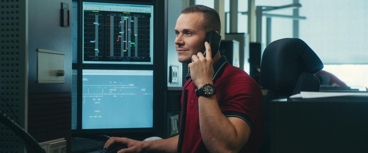 Mann in roten T-Shirt vor Bildschirmen am Telefon.