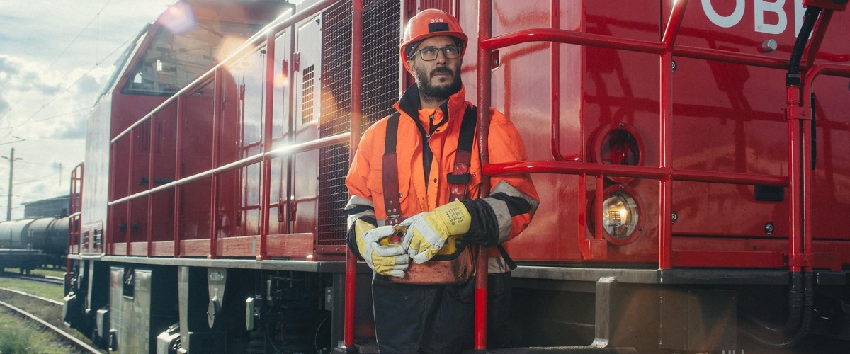 Mann in oranger Uniform auf einem Triebwagen stehend mit Helm und gelben Handschuhe.