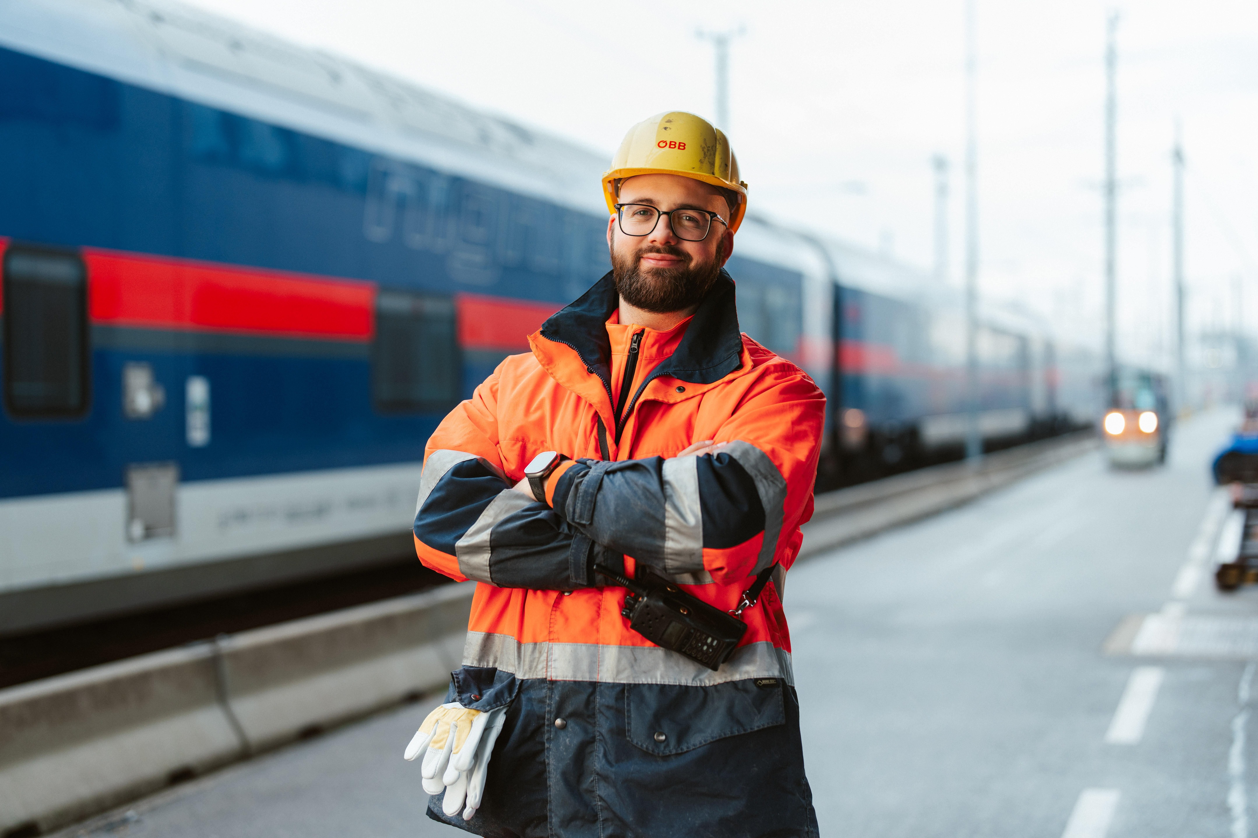 Mann mit orangener Uniform und gelben Helm der eine schwarze Brille trägt. Stehend vor einem blauen Nachtzug