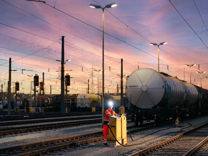 Ein Mann in orangener Uniform im Gleisbereich bei Sonnenuntergang. Der Himmel ist lila verfärbt.