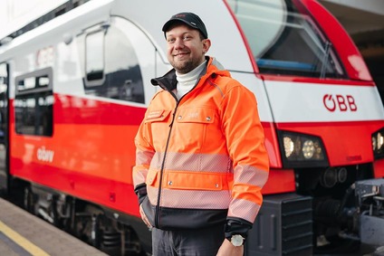 mann mit schwarzer Kappe und orangener Jacke. Stehend vor einem Zug