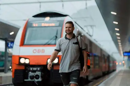 Mann mit grauen Polo und Rucksack umgehängt auf der rechten Seite. Gehend vor einer Lok stehend