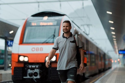 Mann mit grauen Polo und Rucksack umgehängt auf der rechten Seite. Gehend vor einer Lok stehend