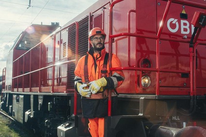 Mann in oranger Uniform auf einem Triebwagen stehend mit Helm und gelben Handschuhe.