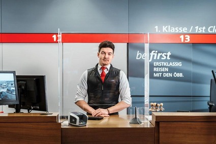 Junger Mann mit roter Krawatte und schwarzen Gilett. Stehend in einem Reisezentrum