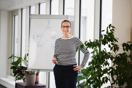 Frau mit gestreiften Shirt und schwarzer Brille. Vor einem Whiteboard stehend mit Stift in der Hand
