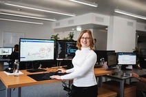 Frau mit schulterlangen braunen Haaren und schwarzer Brille vor dem Bildschirm stehend