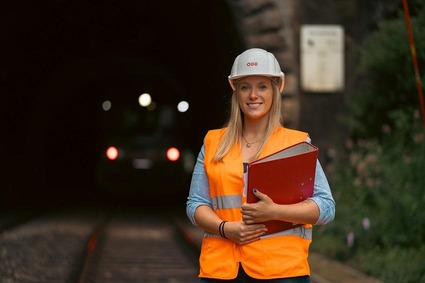 Frau mit blonden langen Haaren und weißen Helm steht vor einem Tunnel mit orangener Warnweste und einer Mappe