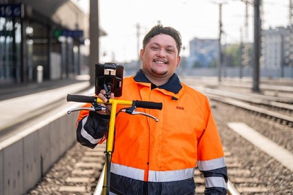 Mann mit kurzen schwarzen Haaren steht lächelnd in orangener Uniform und einen Messgerät auf den Gleisen.