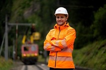Mann mit orangener Uniform und weißen Helm steht mit verschränkten Armen am Gleis.