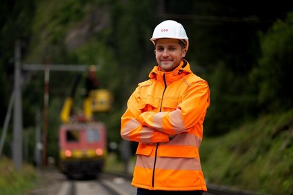 Mann mit orangener Uniform und weißen Helm steht mit verschränkten Armen am Gleis.