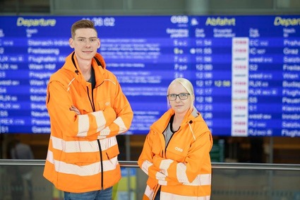 Zwei Mitarbitende stehen in orangener Uniform vor einem Fahrplanbildschirm.