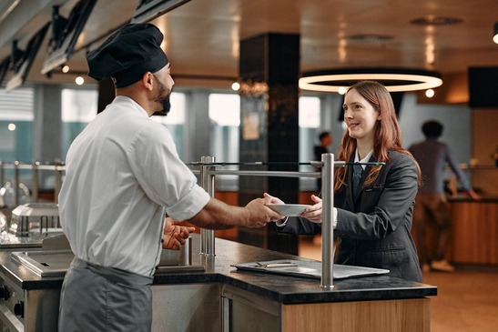 Koch in Uniform übergibt Speise einer jungen Frau mit braunen Haaren in grauer Uniform.