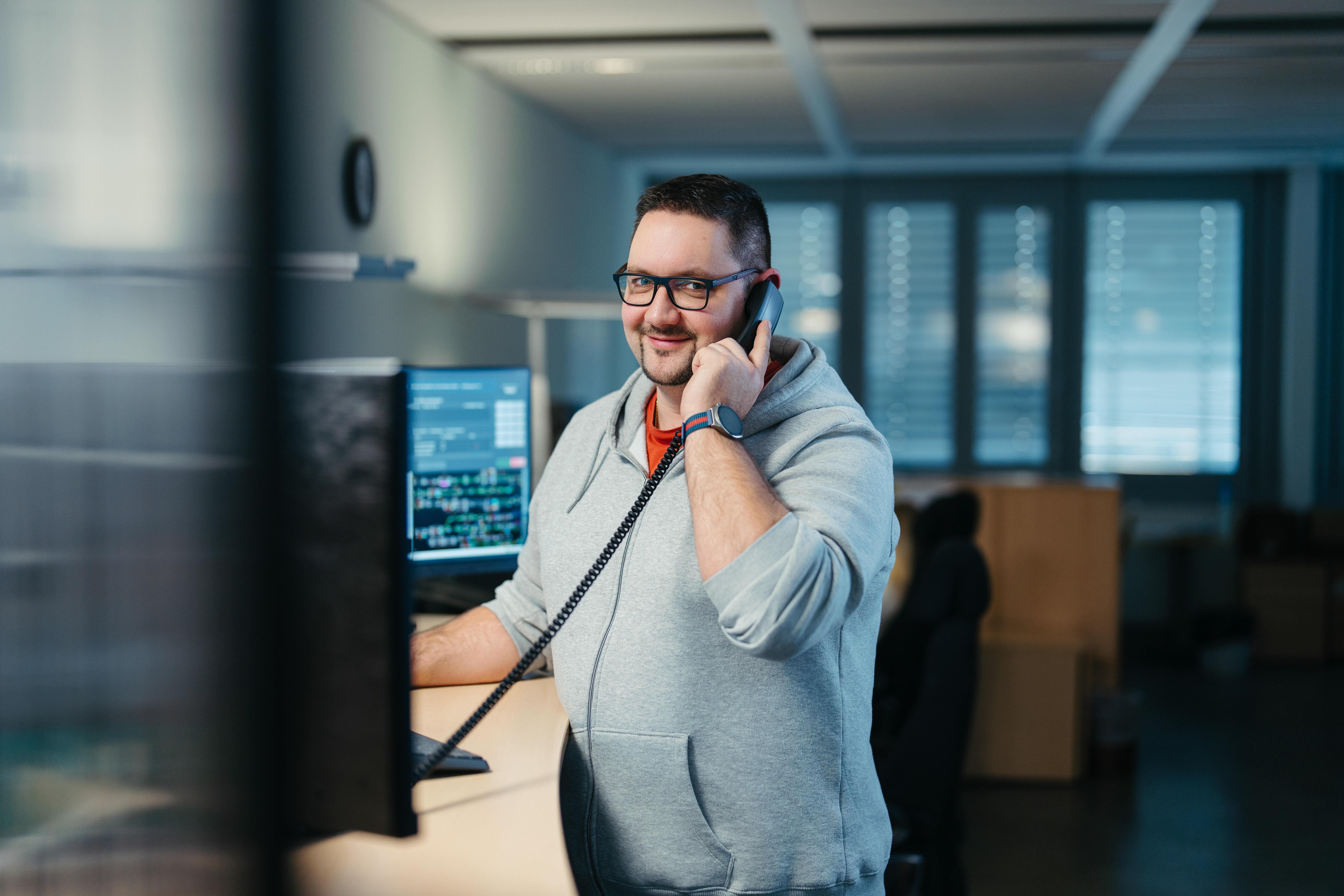 Mann mit Brille vor Bildschirmen stehend und am Telefon.