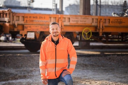 Mann mit kurzen braunen Haaren und orangener Uniform im Gleisbereich stehend.