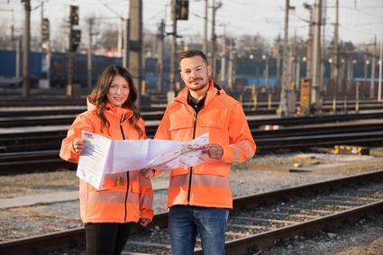 Ein Mann und eine Frau in orangener Uniform im Gleisbereich stehend mit einem Plan in der Hand.