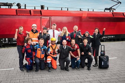 Eine Gruppe Mitarbeitende der ÖBB in verschiedenen Uniformen jubelnd und vor einem Zug stehend