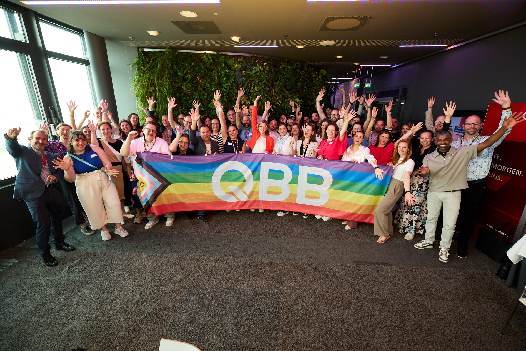 Eine große Gruppe an Mitarbeitende des Regenbogennetzwerkes jubelnd und mit  einer großen Regenbogenflagge stehend mit der Aufschrift QBB.