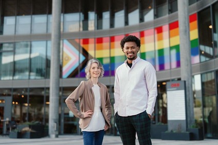 Eine feminin gelesene und eine maskulin gelesene Person mit unterschiedlicher Hautfarbe stehen vor einem modernen Gebäude; hinter ihnen ist eine große Regenbogen- und Progress-Pride-Flagge angebracht