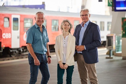 Eine ältere Frau und daneben jeweils ein älterer Mann stehend und lächelnd am Bahnhof.