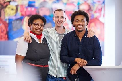 Eine feminin gelesene und zwei maskulin gelesene Person mit unterschiedlicher Hautfarbe stehen vor einem bunten Gemälde.