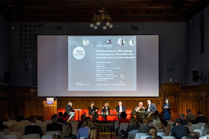 Eine Bühne mit einer großen Leinwand im Hintergrund mit Beteiligten von der Veranstaltung zur Podiumsdiskussion "Fit4Generations. Wie gelingt ausgewogenes Generationsmanament in Unternehmen"