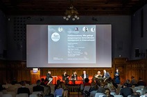 Eine Bühne mit einer großen Leinwand im Hintergrund mit Beteiligten von der Veranstaltung zur Podiumsdiskussion "Fit4Generations. Wie gelingt ausgewogenes Generationsmanament in Unternehmen"