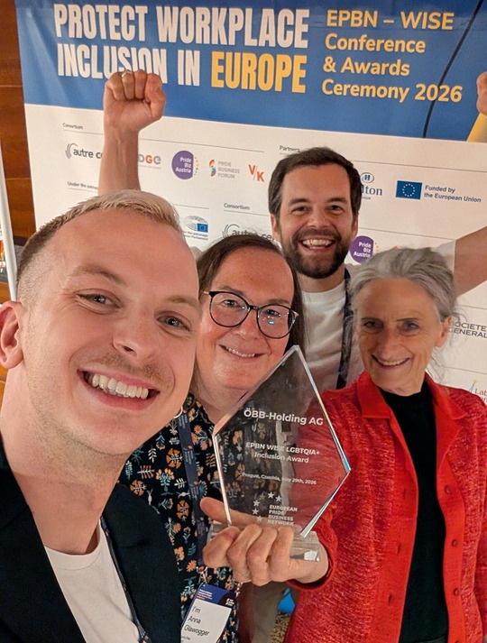 Foto aus der Selfie-Perspektive von vier ÖBB Mitarbeiter:innen, die lachend in die Kamera schauen. Eine Mitarbeiterin hält den EPBN WISE LGBTQIA+ Inclusion Award in die Kamera.