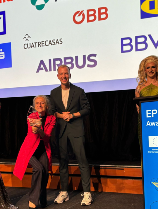 Zwei ÖBB Mitarbeiter:innen auf der Bühne, lachen in die Kamera. Eine Mitarbeiterin hält dabei den EPBN WISE LGBTQIA+ Inclusion Award in die Kamera. Daneben steht ein:e Drag Artist:in am Redner:innenpult.