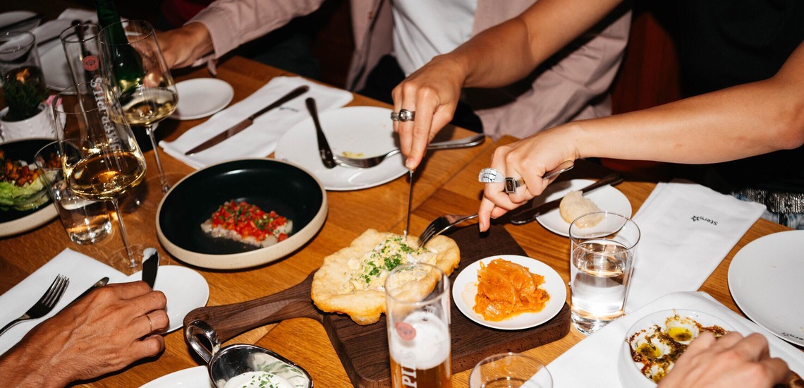 Essen in Wien: Ein Tisch voller Gläser, Teller mit Essen und Gläsern mit Getränken, verschiedene Personen greifen zu. Zu sehen sind nur Hände mit Besteck.