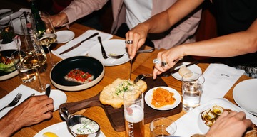 Essen in Wien: Ein Tisch voller Gläser, Teller mit Essen und Gläsern mit Getränken, verschiedene Personen greifen zu. Zu sehen sind nur Hände mit Besteck.