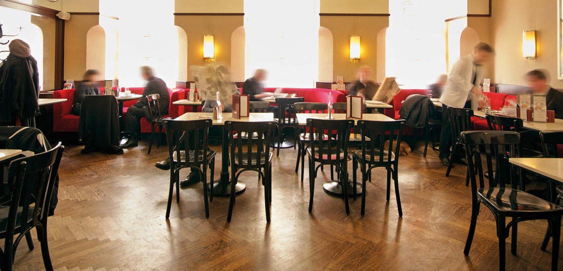 Essen in Wien: Blick auf ein typisches Wiener Kaffeehaus mit kleinen Tischen und schwarzen Sesseln. Dort und da sitzen Gäste, dazwischen sieht man verschwommen Kellner:innen, die zwischen den Tischen gehen. 