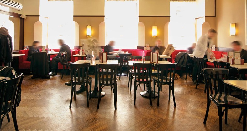 Essen in Wien: Blick auf ein typisches Wiener Kaffeehaus mit kleinen Tischen und schwarzen Sesseln. Dort und da sitzen Gäste, dazwischen sieht man verschwommen Kellner:innen, die zwischen den Tischen gehen. 