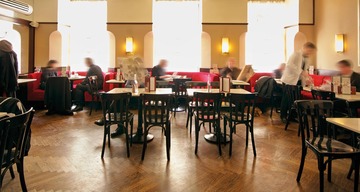 Essen in Wien: Blick auf ein typisches Wiener Kaffeehaus mit kleinen Tischen und schwarzen Sesseln. Dort und da sitzen Gäste, dazwischen sieht man verschwommen Kellner:innen, die zwischen den Tischen gehen. 