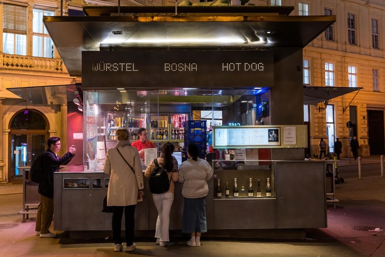 Zu den Wien Sehenswürdigkeiten gehört auch der Würstelstand. Im Bild sieht man Menschen, die sich abends oder nachts vor einem modernen Würstelstand anstellen. 