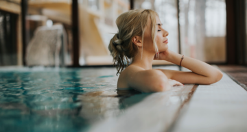 Wellness in Österreich: Eine Frau steht am Beckenrand und hat die Augen genüsslich geschlossen, hinter ihr hellblaues Wasser. 