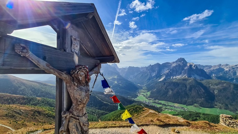 Pustertal: Blick vom Weitwanderweg Via della pace an einem sonnigen Sommertag. Im Hintergrund die schneebedeckten Gipfel, links vorne ein Teil von einem Kreuz mit Jesusfigur. 