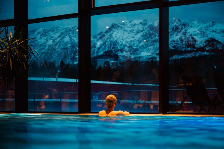 Schladming: Wer in der ThermeGrimming badet hat einen schönen Blick auf die Bergwelt rundum. Im Bild ein Mann im Wasser, der bei einem großen Fenster auf die nächtlichen Berge blickt. 