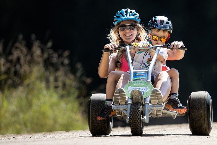 Schladming Mountain Gokart: Eine Frau sitzt zusammen mit einem Kind auf einem Mountain Gokart, beide lachen und fahren gerade bergab. Es ist sonniges Wetter, sie haben kurze Hosen und T-Shirts sowie Helme an. 