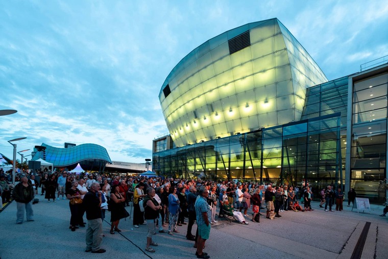 Zu den Sehenswürdigkeiten St. Pölten gehört das Landestheater. Auf dem Bild sieht man es bei Nacht, beleuchtet und mit vielen Passant:innen.