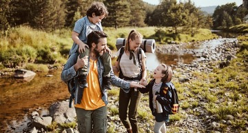 Tagesausflug ab Wien: Eine junge Familie mit Mutter, Vater und zwei Kindern wandert neben einem Back. Die Mutter hat einen Rucksack samt zusammengerollter Sitzunterlage auf dem Rücken. Ein Kind sitzt auf den Schultern des Vaters, ein Kind trägt einen Rucksack. 