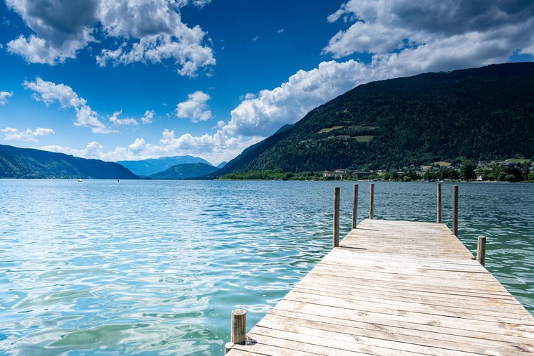 Blick auf den Ossiacher See bei Villach. Ein Steg ragt rechts in den See, am Horizont Berge. 