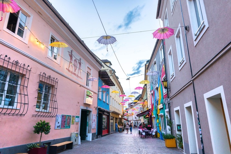 Blick in eine Altstadtgasse in Villach, mit bunten Häusern und Regenschirm-Dekoration. 