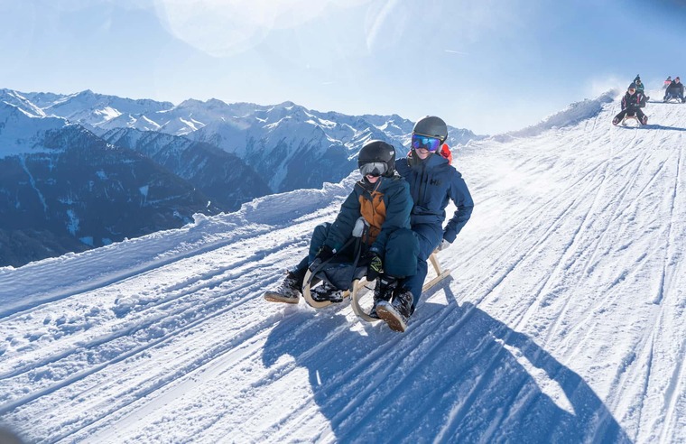 Winterurlaub in Österreich: Auf dem Bild sieht man einen Erwachsenen und ein Kind auf einem Schlitten, auf der Rodelbahn in Bramberg am Wildkogel. Im Hintergrund das Bergpanorama. 