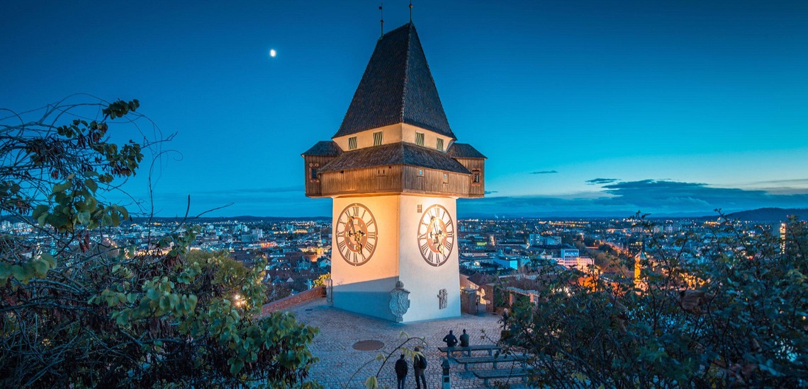 Graz Sehenswürdigkeiten: Blick auf den Grazer Uhrturm und die dahinterliegende Stadt bei Nacht. 