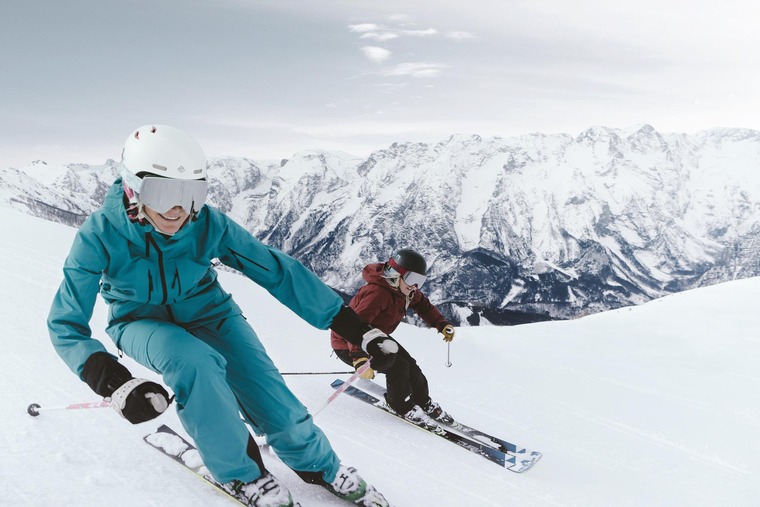 Zug zum Skifahren: Abfahrten in Hinterstoder versprechen pures Skivergnügen. Auf dem Bild zu sehen sind zwei Skifahrerinnen, die einen Hang in Skigebiet sportlich befahren. Im Hintergrund sind die schneebedeckten Berge zu sehen. 