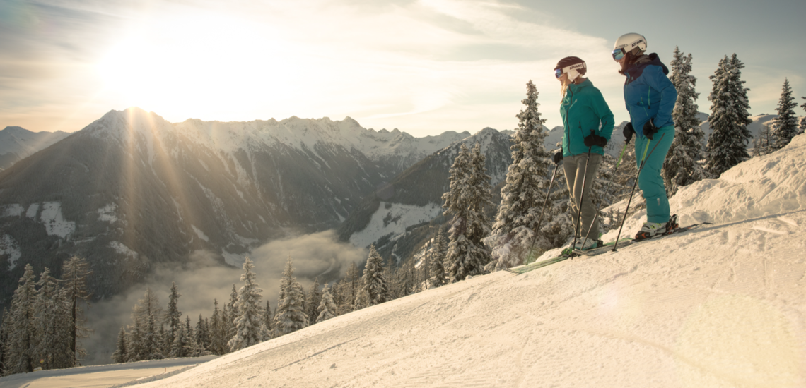 Skiurlaub mit dem Zug in Österreich: 2 Skifahrer:innen stehen auf einem Skihang und blicken in Richtung Tal. Dahinter steht die Sonne über einer Bergkette. 