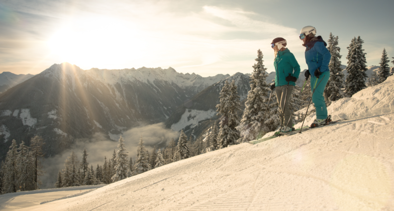 Skiurlaub mit dem Zug in Österreich: 2 Skifahrer:innen stehen auf einem Skihang und blicken in Richtung Tal. Dahinter steht die Sonne über einer Bergkette. 